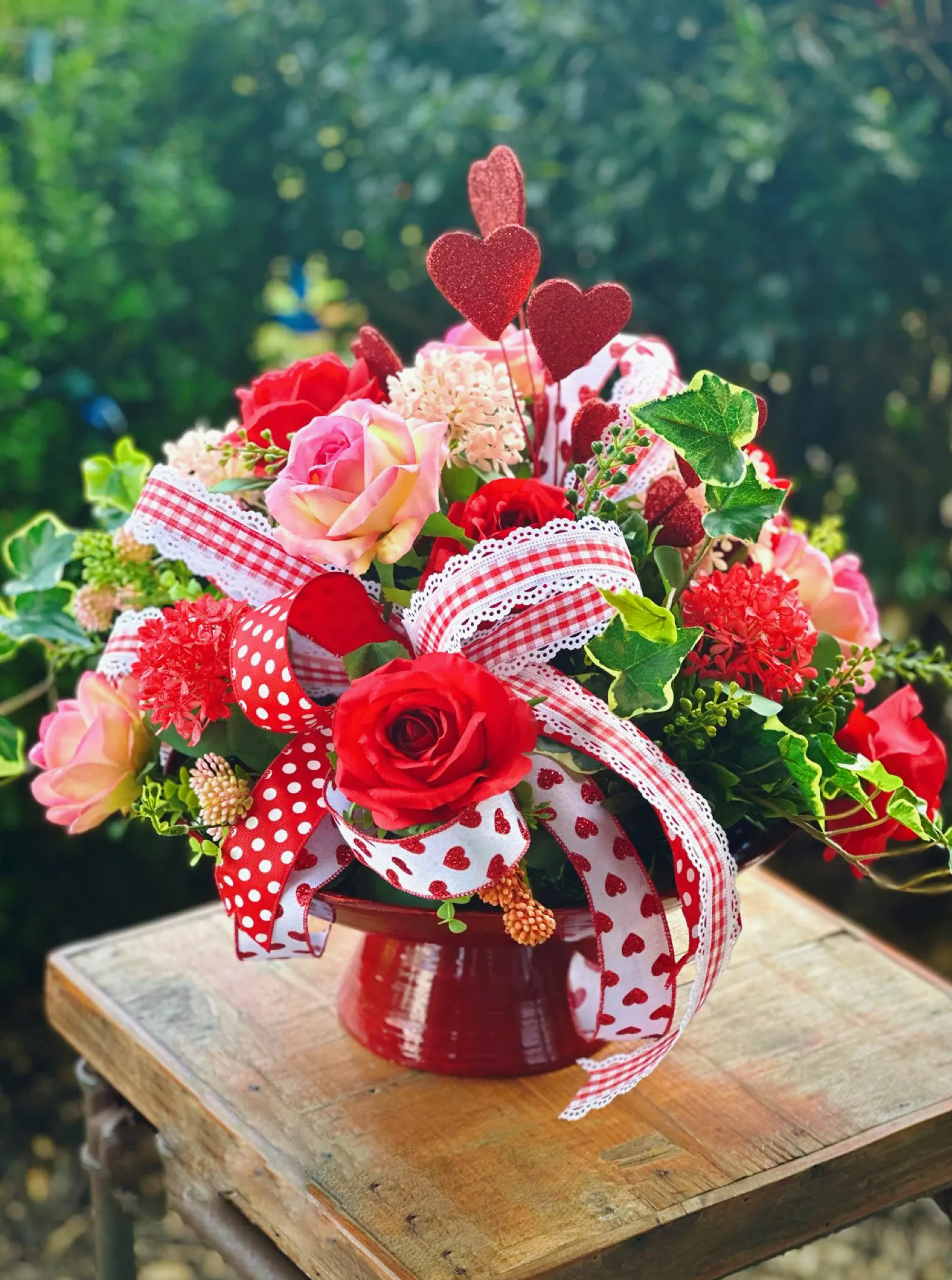 A Valentine's Day arrangement with red and pink roses, heart picks, and gingham ribbon in a red enamel pot